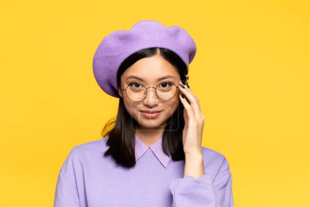 happy asian woman in beret adjusting eyeglasses isolated on yellow 