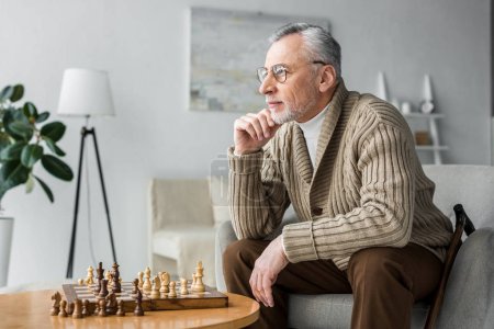 thoughtful retired man in glasses thinking while sitting near chess board at home