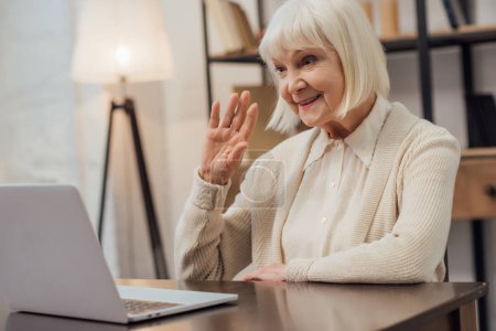 smiling senior woman sitting at computer desk and waving while having video call at home
