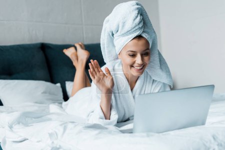 happy woman waving and having video call on laptop while lying on bed