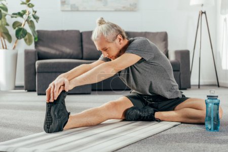 sportive adult man in sportswear sitting and stretching on yoga mat at home