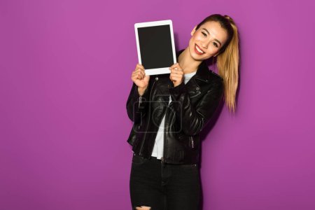 beautiful young woman holding digital tablet and smiling at camera isolated on violet