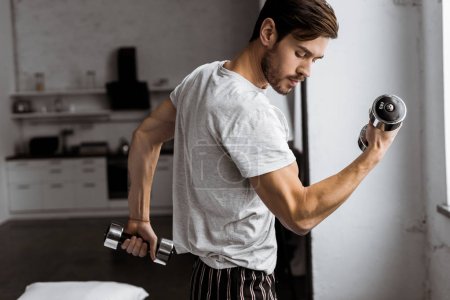 handsome young man in pajamas exercising with dumbbells and looking at biceps at home