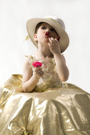Sweet brunette little girl in light goldish dress and white hat making up her lips