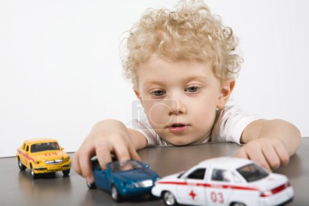 Curly blond boy playing with cars on the gray table