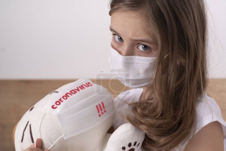 The girl is sad because of the virus.Little sad girl in a white T-shirt in a protective mask plays with a toy.Concept of the fight against coronovirus and other infectious diseases.