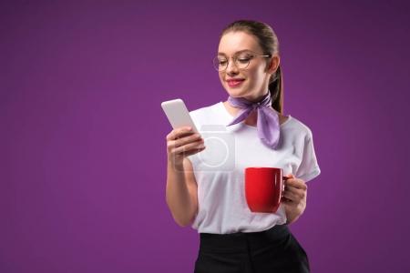 girl with red cup of coffee using smartphone isolated on purple