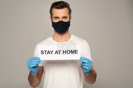 Man in safety mask and latex gloves holding card with stay at home lettering isolated on grey 