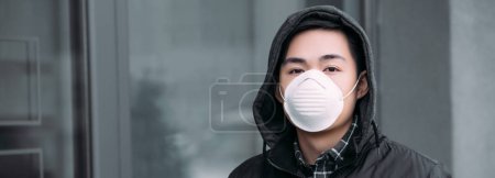 panoramic shot of young asian man in respirator mask looking at camera while standing on street