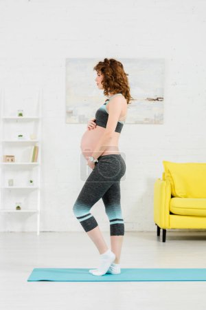 Side view of pregnant woman touching belly while standing on fitness mat at home 