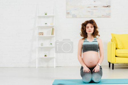 Beautiful pregnant woman with closed eyes sitting on fitness mat in living room
