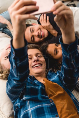 overhead view of multicultural teens taking selfie while lying on bed 