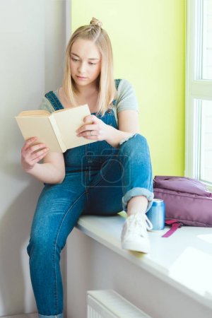 Teenage schoolgirl reading book and sitting on window sill with soda and backpack