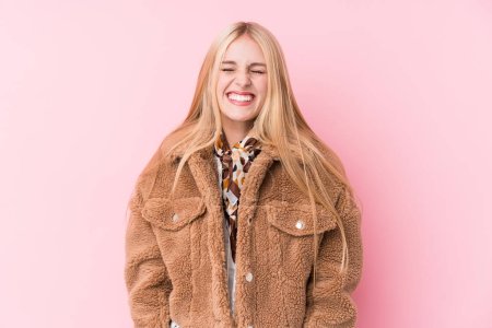 Young blonde woman wearing a coat against a pink background laughs and closes eyes, feels relaxed and happy.