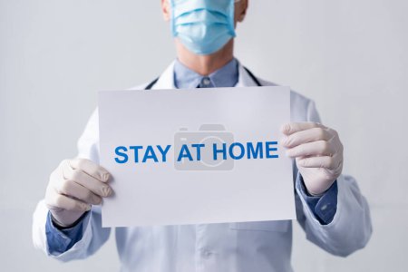 cropped view of middle aged doctor in medical mask holding placard with stay at home lettering on grey