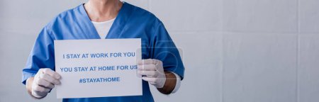 panoramic shot of mature doctor holding placard with i stay at work for you, you stay at home for us lettering on grey