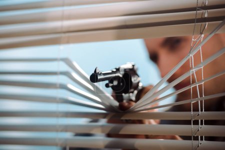 panoramic shot of black gun near window blinds in hand of gangster 