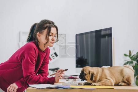 Attractive young girl playing with pug while working on illustrations in home office