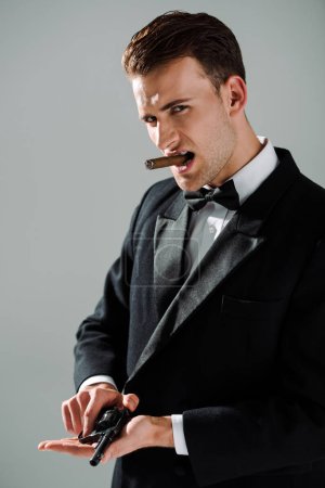 stylish gangster in suit with bow tie holding gun while smoking cigar isolated on grey 