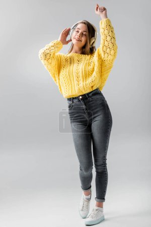 full length view of smiling girl dancing while listening music in wireless headphones with closed eyes on grey background