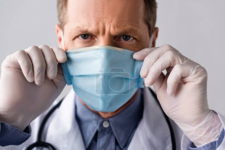 mature doctor touching blue medical mask isolated on grey 
