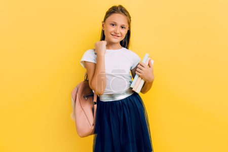 A teenager with a backpack and books, a schoolgirl Girl shows a victory gesture on a yellow background