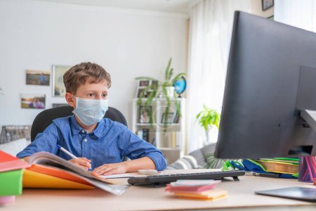 Boy in face mask with computer does his homework during a coronavirus quarantine. concept of online education. Social distance, self- isolation. Distance learning due to virus, flu, epidemic COVID-19