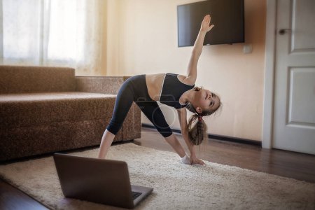 Pretty young girl in sportswear watching online video on laptop and doing fitness exercises at home. Distant training with personal trainer, social distance or self-isolation, online education concept