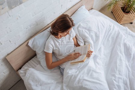 top view of happy woman reading book while chilling in bed 