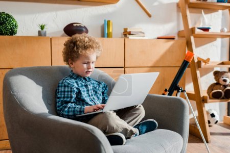 cute kid sitting in armchair and using laptop 