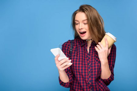 surprised girl with coffee to go using smartphone, isolated on blue