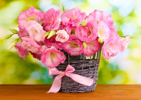 Bouquet of eustoma flowers in wicker vase, on wooden table, on green background
