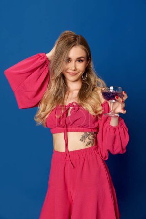 Portrait of charming girl in bright clothes holding cocktail playing with her hair and looking at camera over blue studio background. Concept of fashion, party, beauty, human emotions