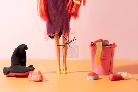 Doll barbie with red hair in a witch costume next to a pink bucket filled with candy.