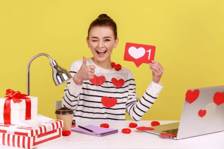 Smiling woman holding like counter template sitting at workplace covered heart sticks, making post in social network, showing thumb up. Indoor studio studio shot isolated on yellow background.